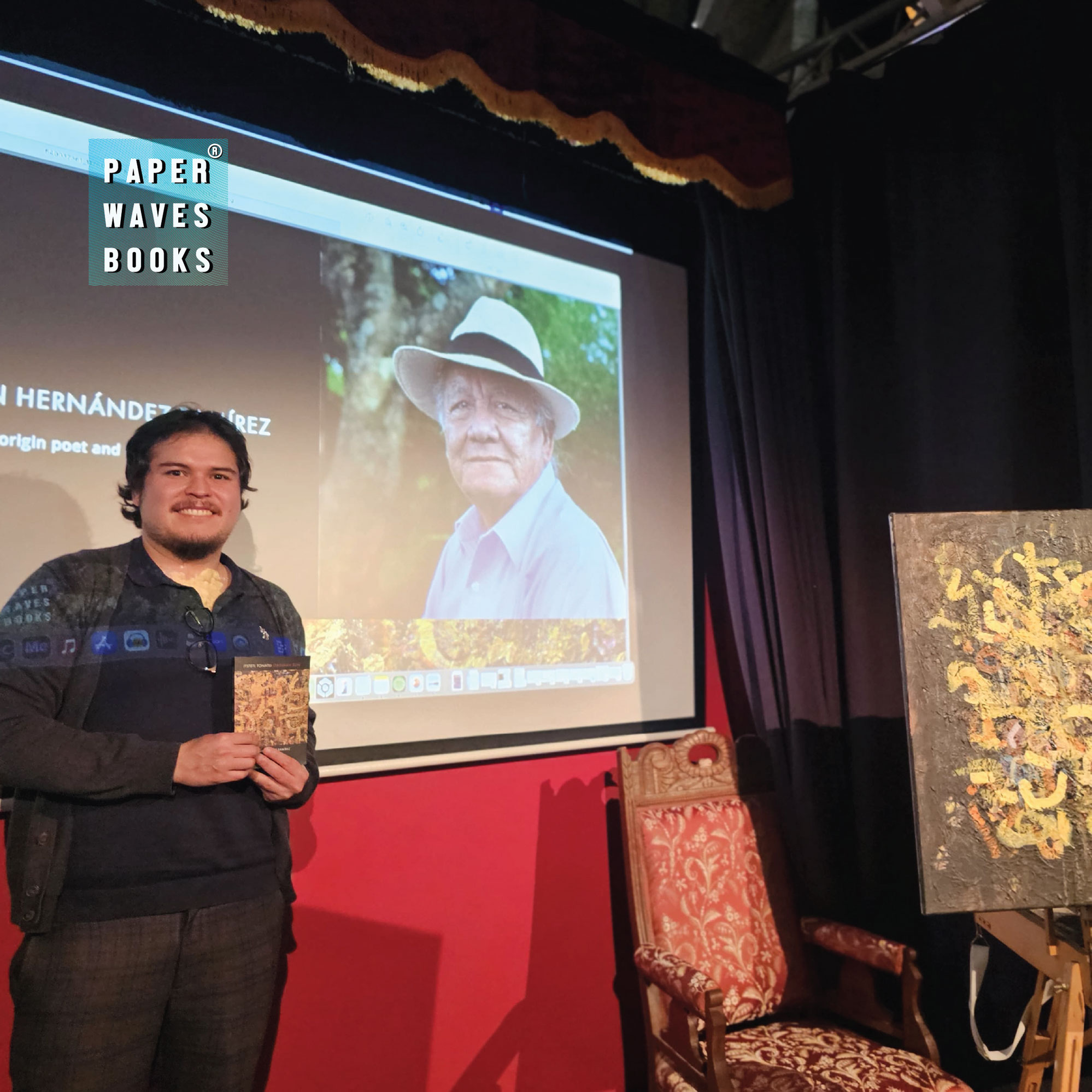 The presenter holding Obsidian Sun beside artwork, with a portrait of the author projected behind him